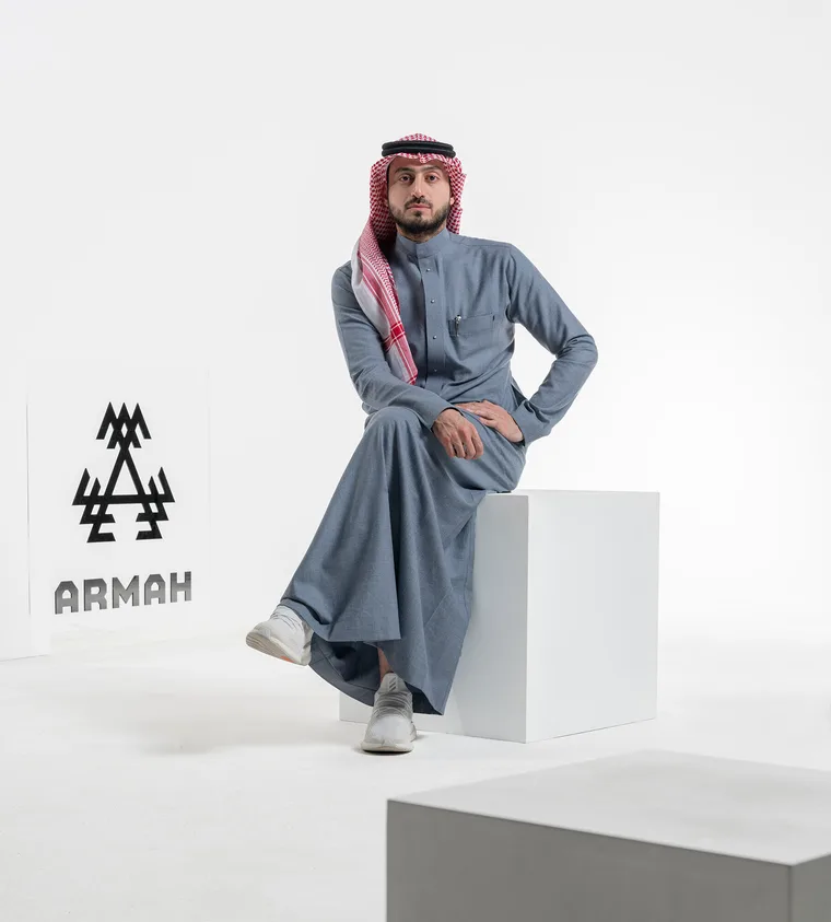 Saudi man wearing a gray thobe and red-white shemagh sitting on a white cube in a clean white studio, with the ARMAH brand logo visible on a sign to his left