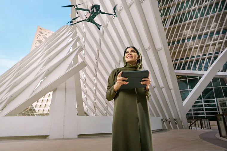 Saudi woman in an olive green abaya holding a tablet and looking up at a flying drone, standing in front of striking modern white geometric architecture under a clear blue sky