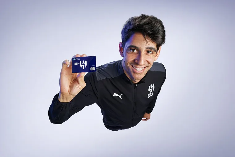 Overhead angle shot of a young Al Hilal football player smiling and holding up a Riyad Bank Mastercard World Elite co-branded card toward the camera, wearing a dark blue Al Hilal Puma training jacket