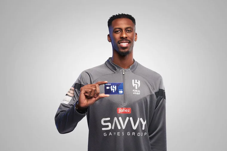 Al Hilal football club player smiling and holding up a Riyad Bank co-branded card, wearing a gray Al Hilal training jersey with Savvy Games Group and Jahez sponsor logos visible