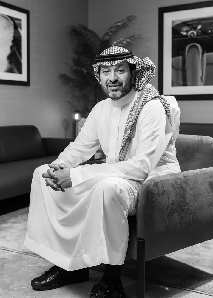 Black and white portrait of a Saudi executive in a white thobe and shemagh seated in a velvet armchair in a luxury lounge, with automotive art prints on the wall, a palm plant, and soft ambient lighting creating a refined atmosphere