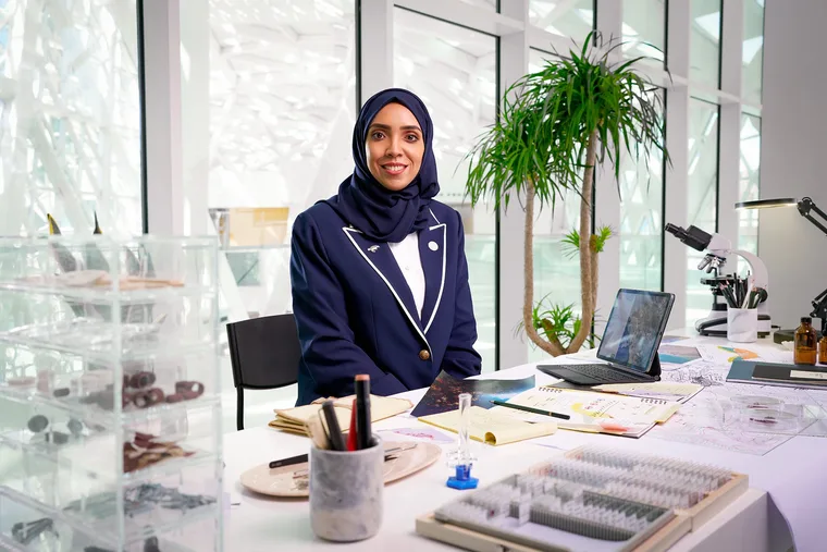 Saudi woman scientist in a navy blue blazer with white trim and dark hijab sitting at a research lab desk surrounded by laboratory equipment, specimen slides, a microscope, a tablet, and notes, with a bright modern architectural glass structure visible through the windows behind her