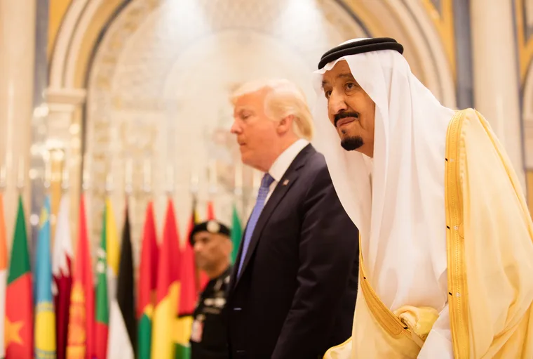 Two world leaders at a formal diplomatic summit walking past a row of international flags in an ornate hall, with a security officer visible in the background