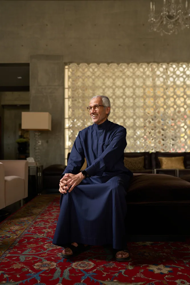 Distinguished senior Saudi man wearing a navy blue thobe and glasses, sitting on a bench in a modern lounge with concrete walls, an illuminated Islamic geometric screen, a crystal chandelier, and a red ornate carpet, smiling warmly to the side