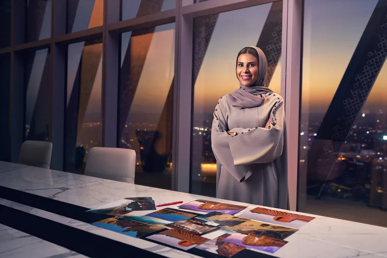Saudi businesswoman in a mauve abaya and hijab standing confidently with arms crossed in a high-rise office at sunset, with architectural photos spread on a marble conference table and a dramatic city skyline glowing through floor-to-ceiling geometric windows