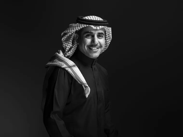 Black and white studio portrait of a smiling Saudi man wearing a dark thobe and a shemagh draped casually over his shoulder, photographed against a dark gradient background with dramatic side lighting
