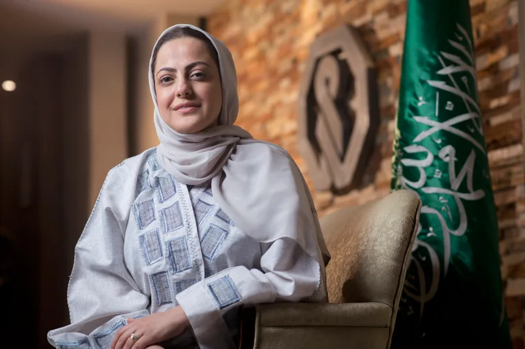 Saudi woman in a white embroidered abaya and beige hijab seated in an upholstered chair in a formal office setting, with the Saudi Arabian flag and a corporate emblem visible in the background against a stone wall